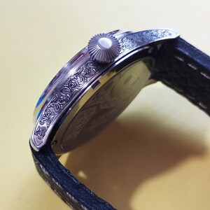 May include: A silver wristwatch with a black leather band. The watch has an intricate engraved design on the side of the case.