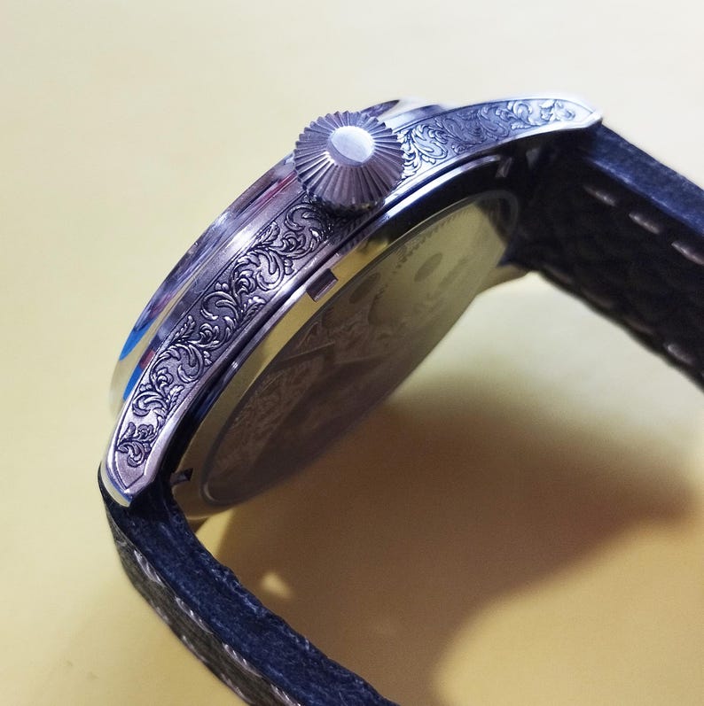 May include: A silver wristwatch with a black leather band. The watch has an intricate engraved design on the side of the case.