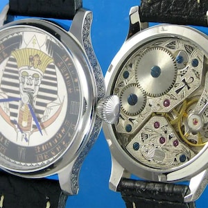 May include: A silver pocket watch with a black leather band. The face of the watch features a detailed illustration of an Egyptian pharaoh's head. The back of the watch shows the intricate inner workings of the watch.