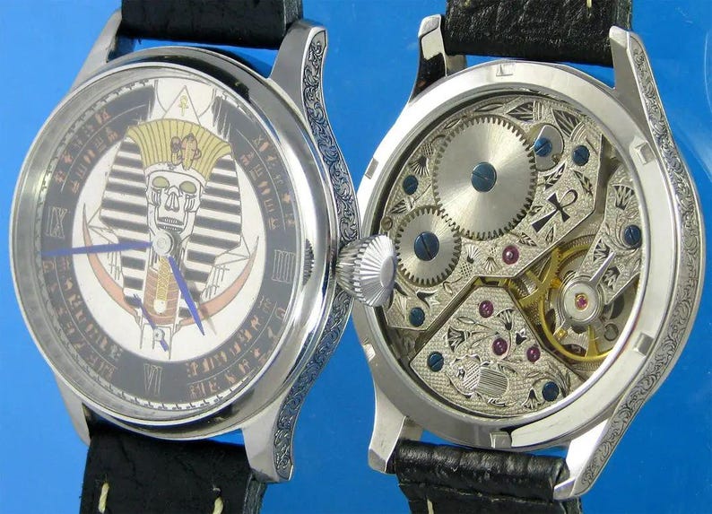 May include: A silver pocket watch with a black leather band. The face of the watch features a detailed illustration of an Egyptian pharaoh's head. The back of the watch shows the intricate inner workings of the watch.