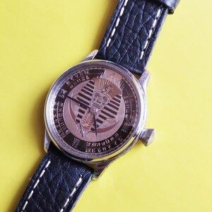 May include: A silver wristwatch with a black leather band. The watch face features a gold-toned design of an Egyptian pharaoh's head with a cobra.