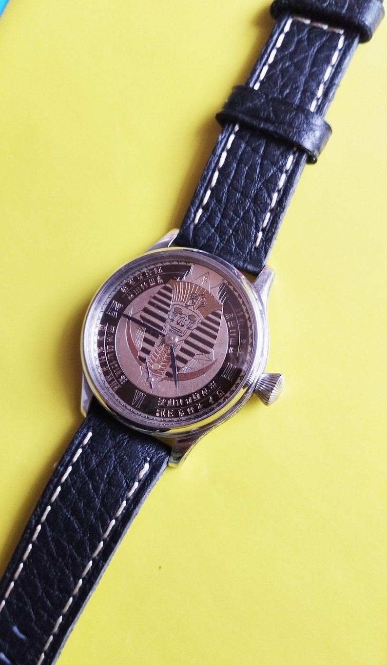 May include: A silver wristwatch with a black leather band. The watch face features a gold-toned design of an Egyptian pharaoh's head with a cobra.