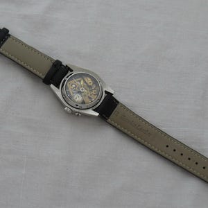 May include: A silver-toned wristwatch with a black leather band and a transparent face revealing the inner workings. The watch has a round face and a buckle clasp. The band is black on the top and light gray on the underside.