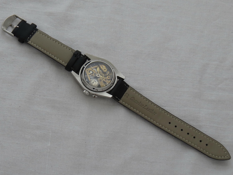 May include: A silver-toned wristwatch with a black leather band and a transparent face revealing the inner workings. The watch has a round face and a buckle clasp. The band is black on the top and light gray on the underside.