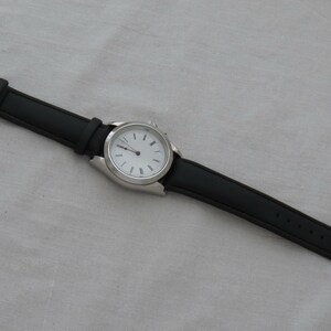 May include: A wristwatch with a black leather band and a silver-colored case. The watch face is white with black hour markers and hands. The band is secured with a silver buckle.