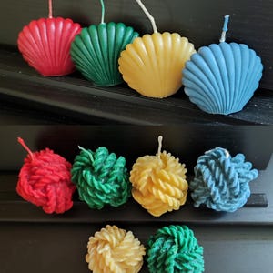 May include: Assortment of colorful candles in two different styles.  The first row features four shell-shaped candles in red, green, yellow, and blue. The second row features six round candles in red, green, yellow, and blue, with a twisted rope design.
