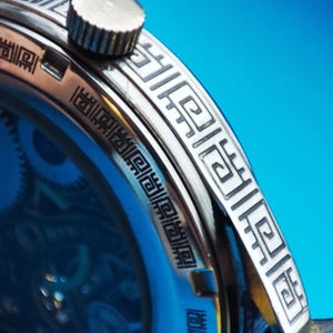 May include: Close-up of a silver watch with a black face and a black leather band. The watch has a decorative pattern around the edge of the face.