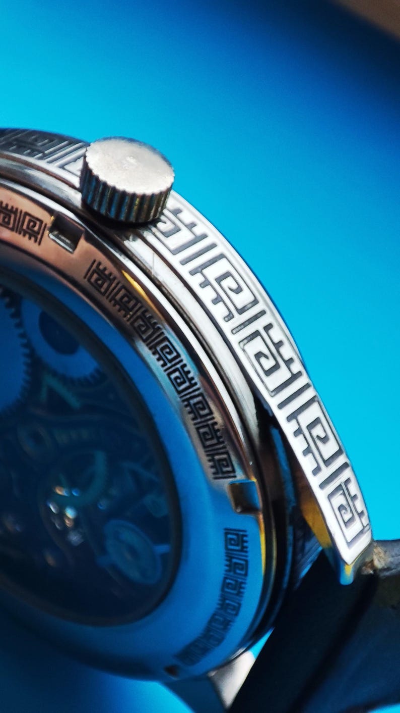 May include: Close-up of a silver watch with a black face and a black leather band. The watch has a decorative pattern around the edge of the face.