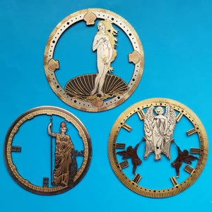 Peut inclure: Trois cadrans de montre décoratifs avec des thèmes d'art classique. L'un représente Vénus sortant d'une coquille, un autre une figure avec une lance, et le troisième un ange avec deux figures. Chacun est doré avec des détails complexes.