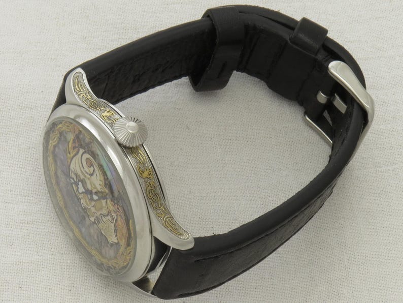 May include: A silver pocket watch with a black leather band. The watch face has a detailed, colorful design with gold accents.