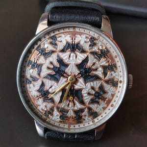 May include: A silver wristwatch with a black leather band. The watch face features a detailed pattern of black, silver, and copper-colored bat-like figures. The hands are gold-colored.