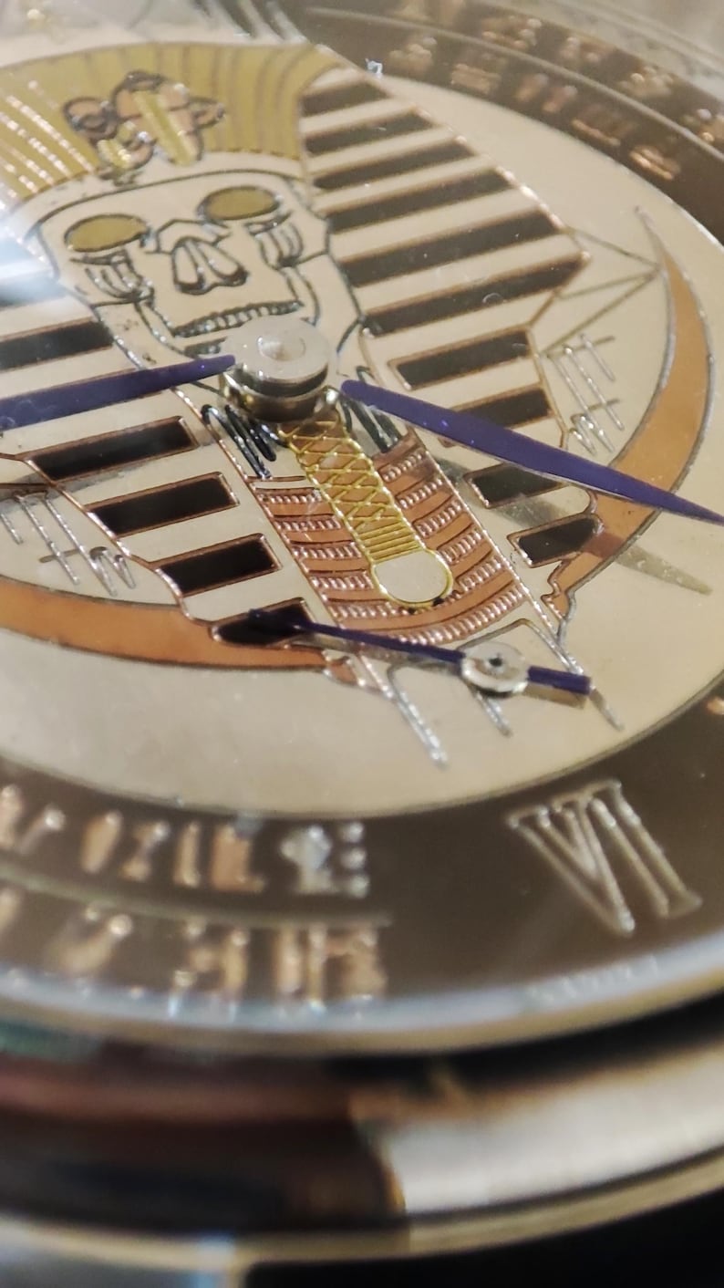 May include: Close-up of a watch face with a gold and black design. The face features a skull with a pharaoh's headdress and Roman numerals around the edge.