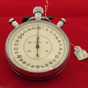 May include: A vintage stopwatch with a silver casing and a cream-colored face. The face features black numerals and a red second hand. The stopwatch has a winding crown and two side buttons. A small tag is attached. The stopwatch is on a red background.