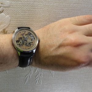 May include: A silver wristwatch with a black leather band. The watch face has a skeleton design with a gold-toned background and intricate details.