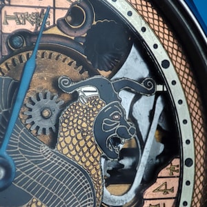 May include: Close-up of a watch face with a black and gold design. The watch face features a detailed illustration of a mythical creature with scales and a fierce expression. The watch face also includes Roman numerals and a copper-colored border.