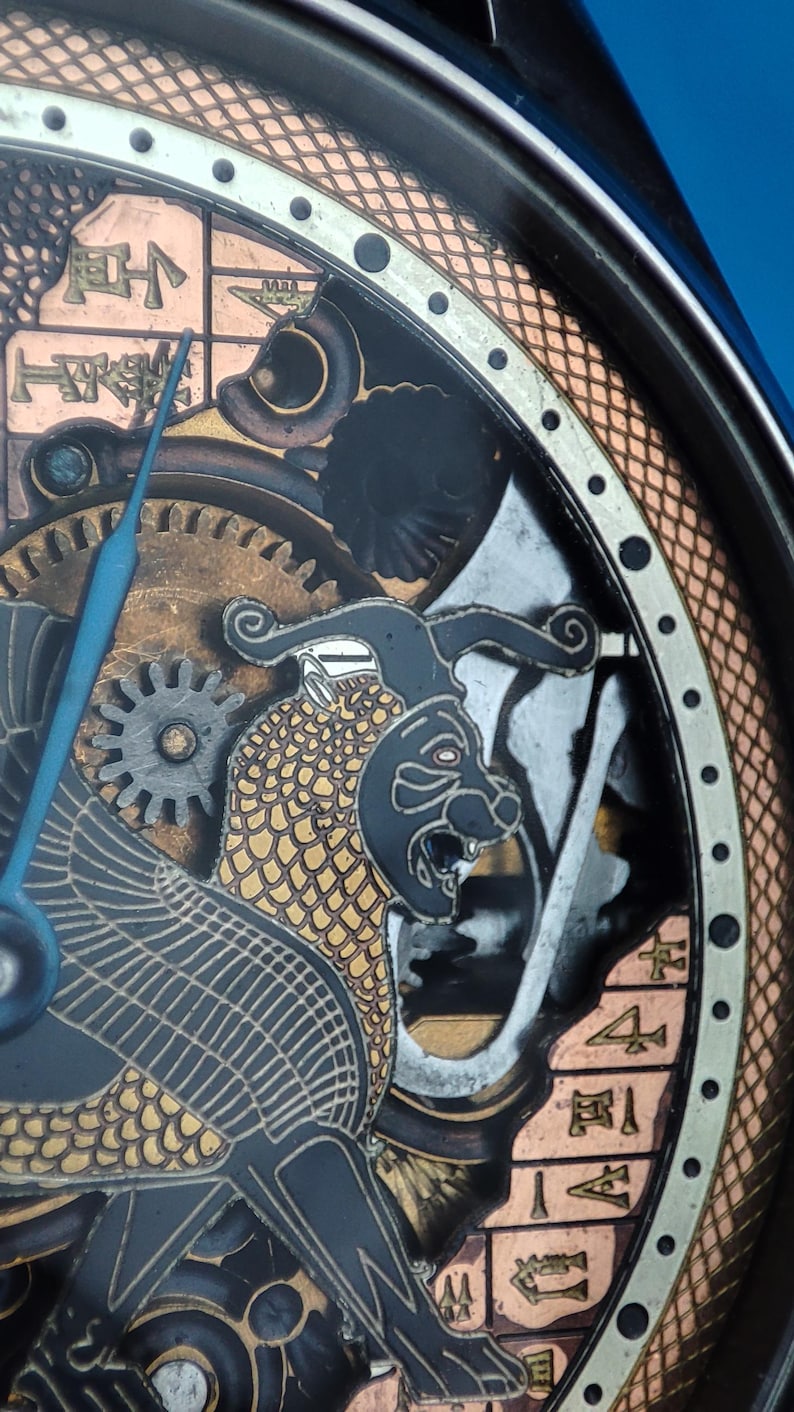 May include: Close-up of a watch face with a black and gold design. The watch face features a detailed illustration of a mythical creature with scales and a fierce expression. The watch face also includes Roman numerals and a copper-colored border.