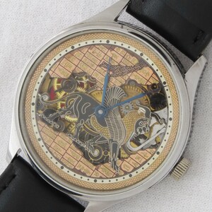May include: A silver-toned wristwatch with a black leather band. The watch face features a gold-toned background with a detailed, intricate design of a mythical creature and gears. The watch face is surrounded by a black ring with white dots.