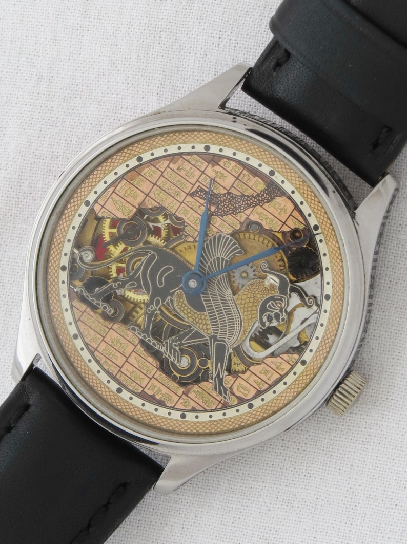 May include: A silver-toned wristwatch with a black leather band. The watch face features a gold-toned background with a detailed, intricate design of a mythical creature and gears. The watch face is surrounded by a black ring with white dots.