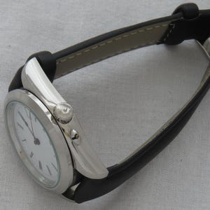 May include: A silver-toned wristwatch with a white face and black hands. The watch has a black leather band with a silver buckle. The watch face displays Roman numerals. The watch is on a light gray surface.