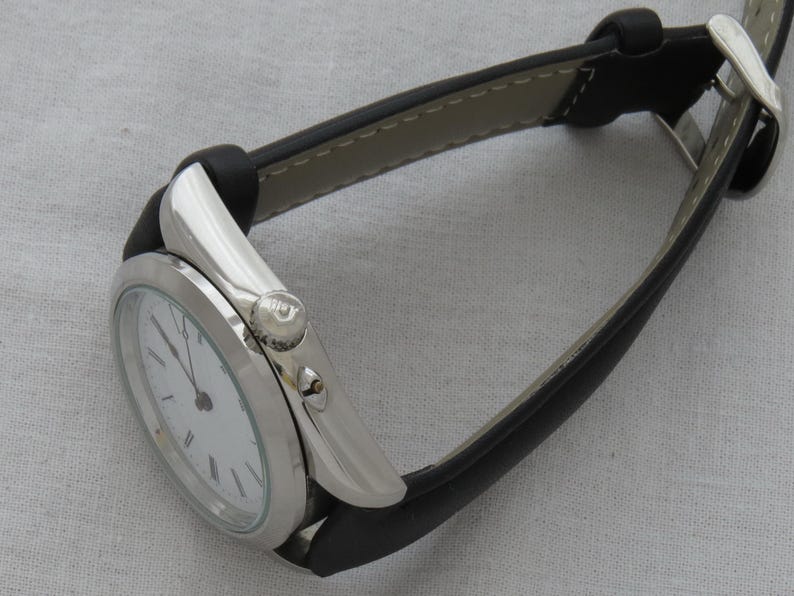 May include: A silver-toned wristwatch with a white face and black hands. The watch has a black leather band with a silver buckle. The watch face displays Roman numerals. The watch is on a light gray surface.