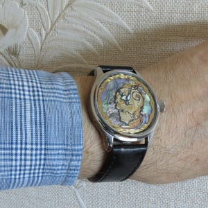 May include: A silver wristwatch with a black leather band. The watch face is round and features a gold-colored design with a mother-of-pearl background.