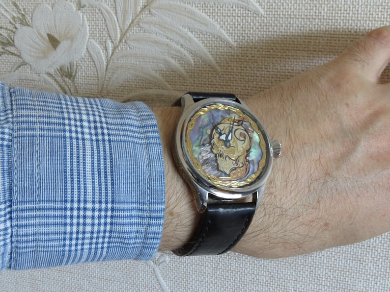 May include: A silver wristwatch with a black leather band. The watch face is round and features a gold-colored design with a mother-of-pearl background.