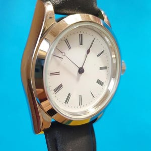 May include: A silver-toned wristwatch with a white face and black Roman numerals. The watch has black hands and a black leather band. The watch face is round and the band is attached to the watch case.