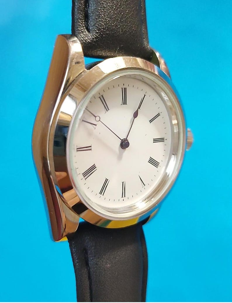 May include: A silver-toned wristwatch with a white face and black Roman numerals. The watch has black hands and a black leather band. The watch face is round and the band is attached to the watch case.