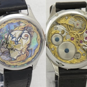 May include: A silver wristwatch with a black leather band. The watch face features a skull design with an abalone shell background. The back of the watch shows the intricate mechanical workings.