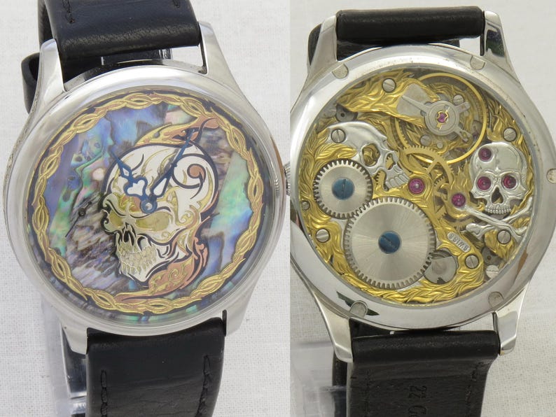 May include: A silver wristwatch with a black leather band. The watch face features a skull design with an abalone shell background. The back of the watch shows the intricate mechanical workings.