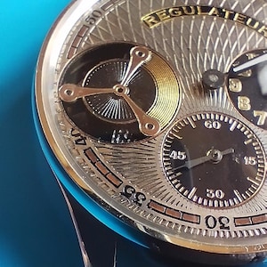 以下が含まれることがあります： 金色文字盤と「Regulateur」という文字が書かれた銀色と黒色の腕時計。黒い革バンドが付いている。