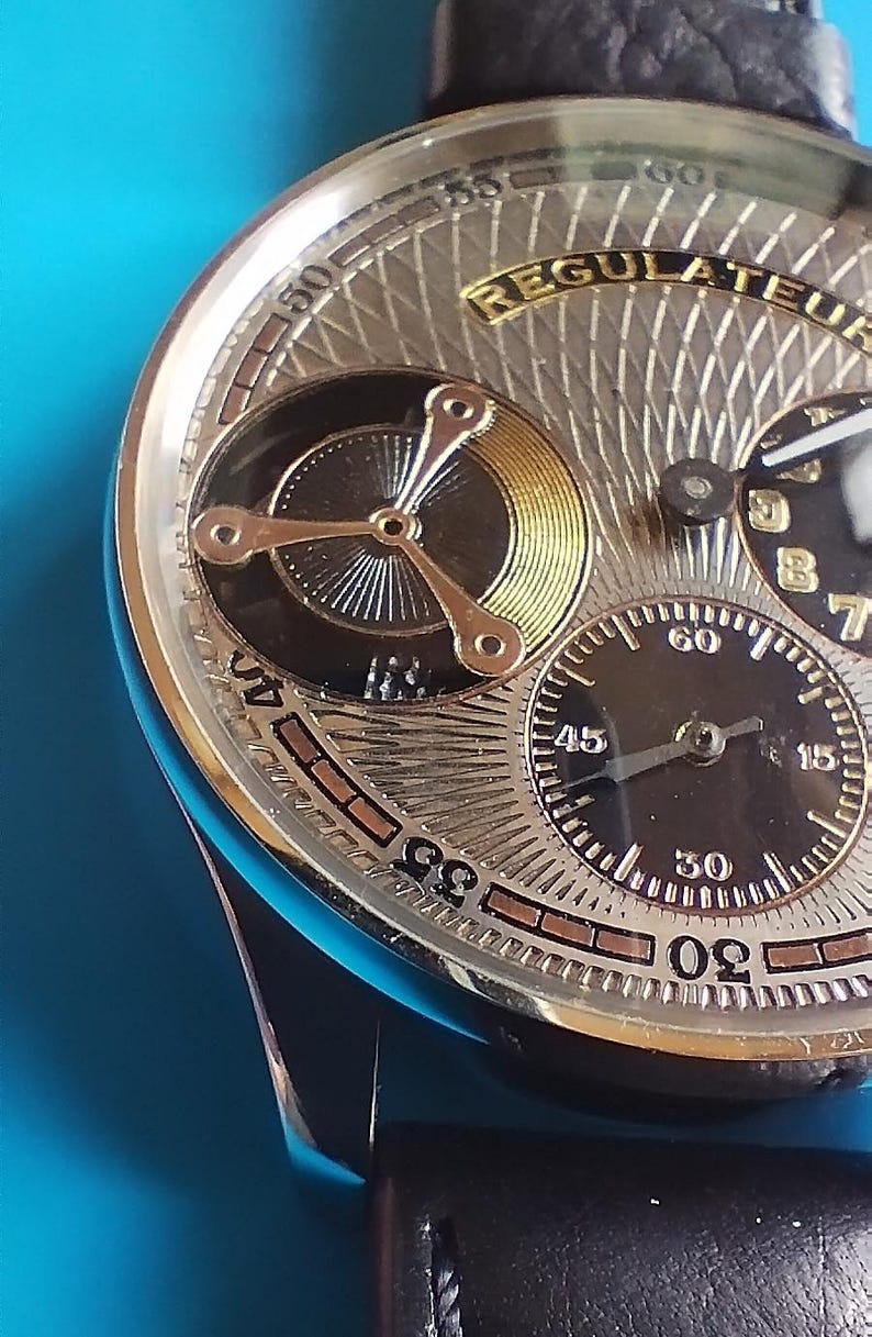 以下が含まれることがあります： 金色文字盤と「Regulateur」という文字が書かれた銀色と黒色の腕時計。黒い革バンドが付いている。