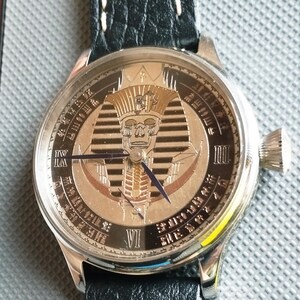 May include: A silver wristwatch with a black leather band. The watch face features a gold and silver design of an Egyptian pharaoh's head with a skull face. The watch face also has Roman numeral markings.