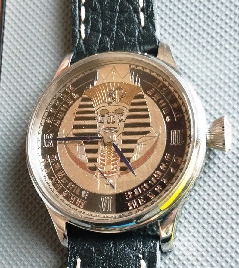 May include: A silver wristwatch with a black leather band. The watch face features a gold and silver design of an Egyptian pharaoh's head with a skull face. The watch face also has Roman numeral markings.
