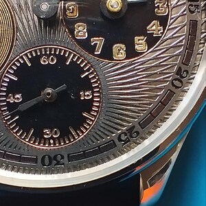 以下が含まれることがあります： 黒の文字盤と金のアクセントが付いた銀色の腕時計。時計には3つの文字盤があり、1つはローマ数字、1つはアラビア数字、1つは0から60までの数字が書かれています。時計は黒い革のストラップに付いています。