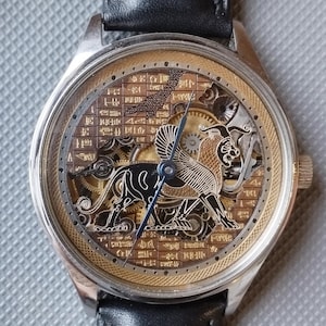 May include: A silver and gold skeleton watch with a black leather band. The watch face features a detailed engraving of a mythical creature with wings and a lion's head.