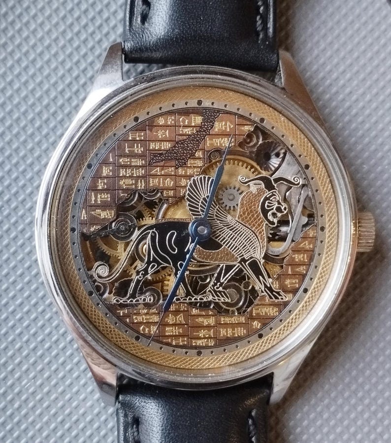 May include: A silver and gold skeleton watch with a black leather band. The watch face features a detailed engraving of a mythical creature with wings and a lion's head.