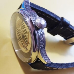 May include: A silver wristwatch with a black leather band. The watch face has a white and black striped design with a gold-colored image in the center. The watch case has an intricate floral design.