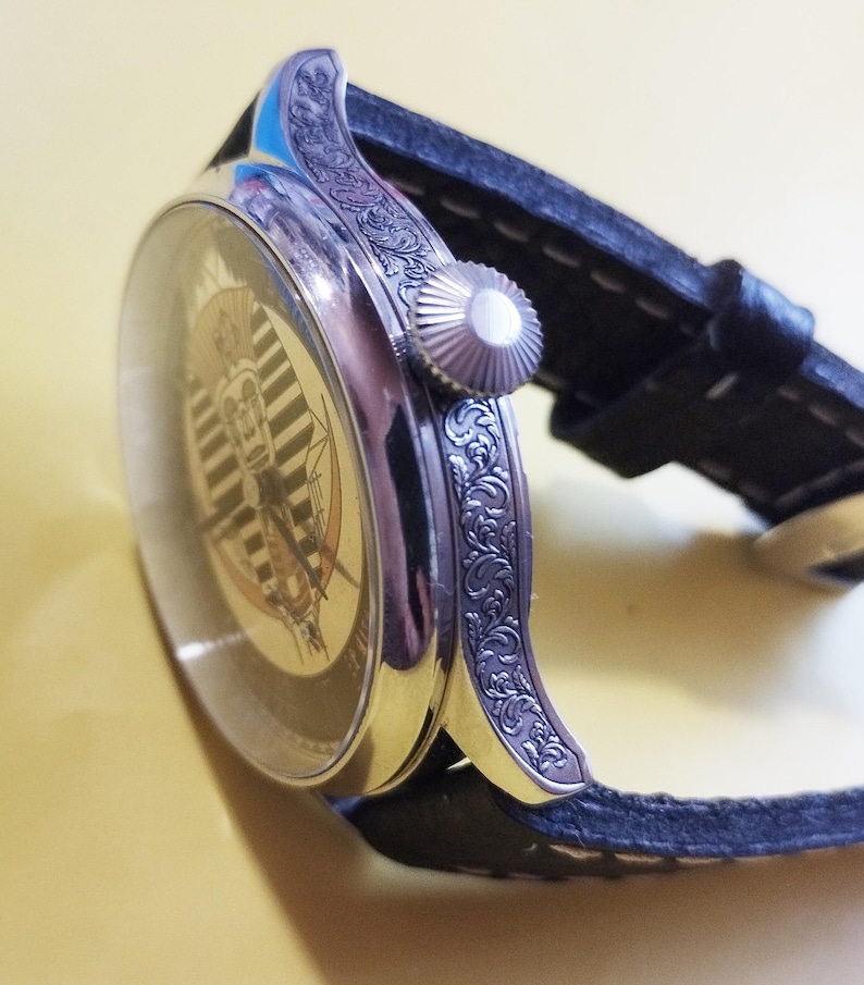 May include: A silver wristwatch with a black leather band. The watch face has a white and black striped design with a gold-colored image in the center. The watch case has an intricate floral design.