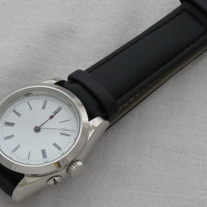 May include: A silver wristwatch with a white face and black Roman numeral hour markers. The watch has black hands and a black leather band. The watch case is approximately 1.5 inches in diameter.