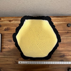 May include: A yellow and black crocheted hexagonal pillow with a black pom-pom trim. The pillow is sitting on a wooden surface.