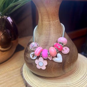 May include: A silver charm bracelet with pink and white charms, including a flower, a heart, a cross, and a smiley face.