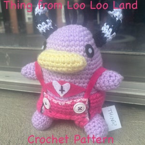May include: A crocheted purple and pink plush toy with black and white horns, a yellow beak, and a pink heart with a cross on its chest. The toy is wearing a pink dress with a white lace trim and a button. The toy is labelled "THING?" and is titled "Thing from Loo Loo Land Crochet Pattern".