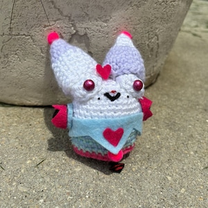 May include: A white crocheted bunny with pink accents and a blue heart on its chest. The bunny has pink eyes and a black smile.