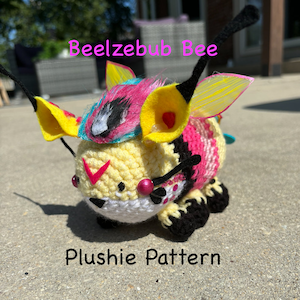 Puede incluir: Un juguete de peluche de ganchillo en forma de abeja con detalles en amarillo, rosa, negro y blanco. La abeja tiene una cabeza grande con una cara rosa y blanca y un cuerpo a rayas blanco y negro. La abeja tiene grandes alas amarillas con rayas negras. La abeja está sentada sobre una superficie gris. El texto "Beelzebub Bee" está escrito encima de la abeja y el texto "Plushie Pattern" está escrito debajo de la abeja.