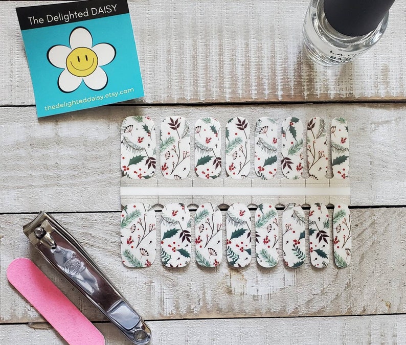 May include: Nail wraps with a white background, featuring a pattern of green leaves, red berries, and brown branches. A bottle of clear nail polish, nail clippers, and a pink nail file are also visible. The Delighted Daisy etsy.com logo is in the top left.