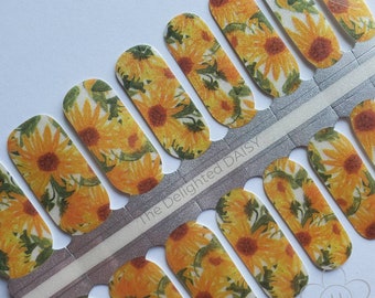 May include: A sheet of nail polish wraps with a sunflower design. The wraps are arranged in a row and feature a white background with yellow sunflowers and green leaves. The text "The Delighted Daisy" is printed on the sheet.