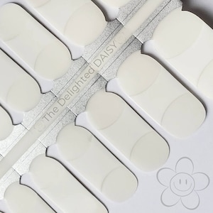 Pode incluir: Close-up de uma folha de tiras de verniz de unhas branco sujo. As tiras são retangulares com extremidades arredondadas e um design curvo. O texto "The Delighted Daisy" está impresso na embalagem. Um gráfico de flor sorridente está no canto.