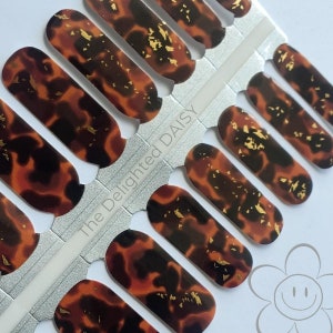 Op de afbeelding: Een vel nagellak wraps met een schildpadpatroon en gouden vlokken. De wraps zijn in een rij op een witte achtergrond gerangschikt. De tekst "The Delighted Daisy" is op het vel gedrukt.