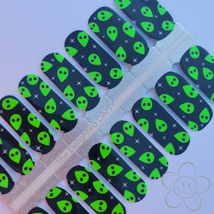 May include: Nail wraps featuring a repeating pattern of green alien faces and white stars on a black background. The nail wraps are on a silver backing with the text "The Delighted Daisy".